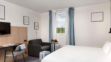 A hotel room with a bed, a desk with a chair, a television, and a small table with a cat.
