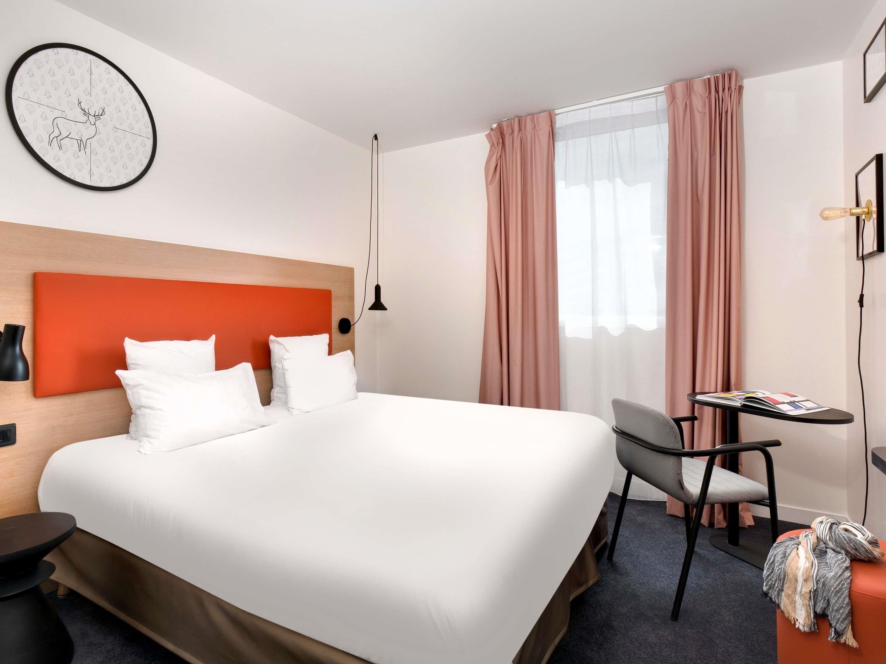 A hotel room with a large bed, a desk, a chair, a clock on the wall, and a window with curtains.