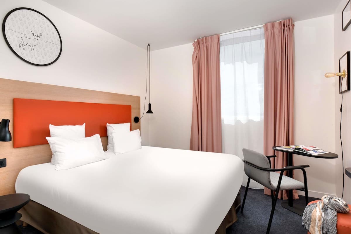 Une chambre d’hôtel avec un grand lit, un bureau, une chaise, une horloge au mur et une fenêtre avec des rideaux.
