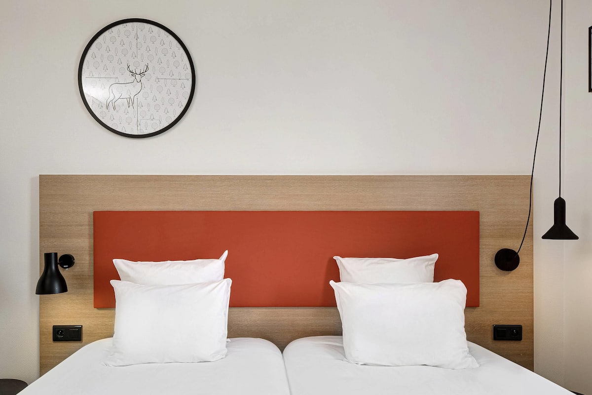 Un lit double avec une literie blanche, une tête de lit en bois, un panneau décoratif rouge, une horloge murale, un lampadaire noir et une plaque d’interrupteur noire.