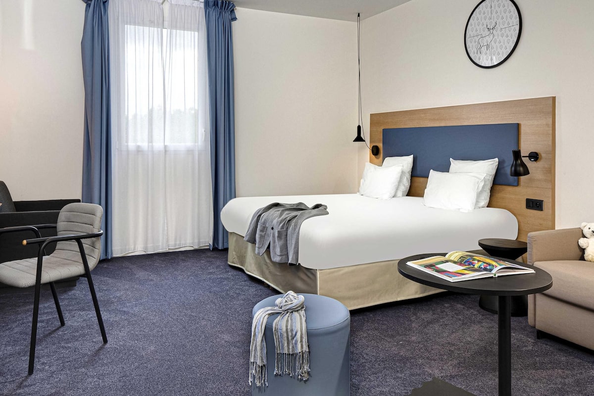 Une chambre d’hôtel comprenant un lit, un bureau, une chaise, une petite table, un canapé, une horloge au mur et une fenêtre avec des rideaux.