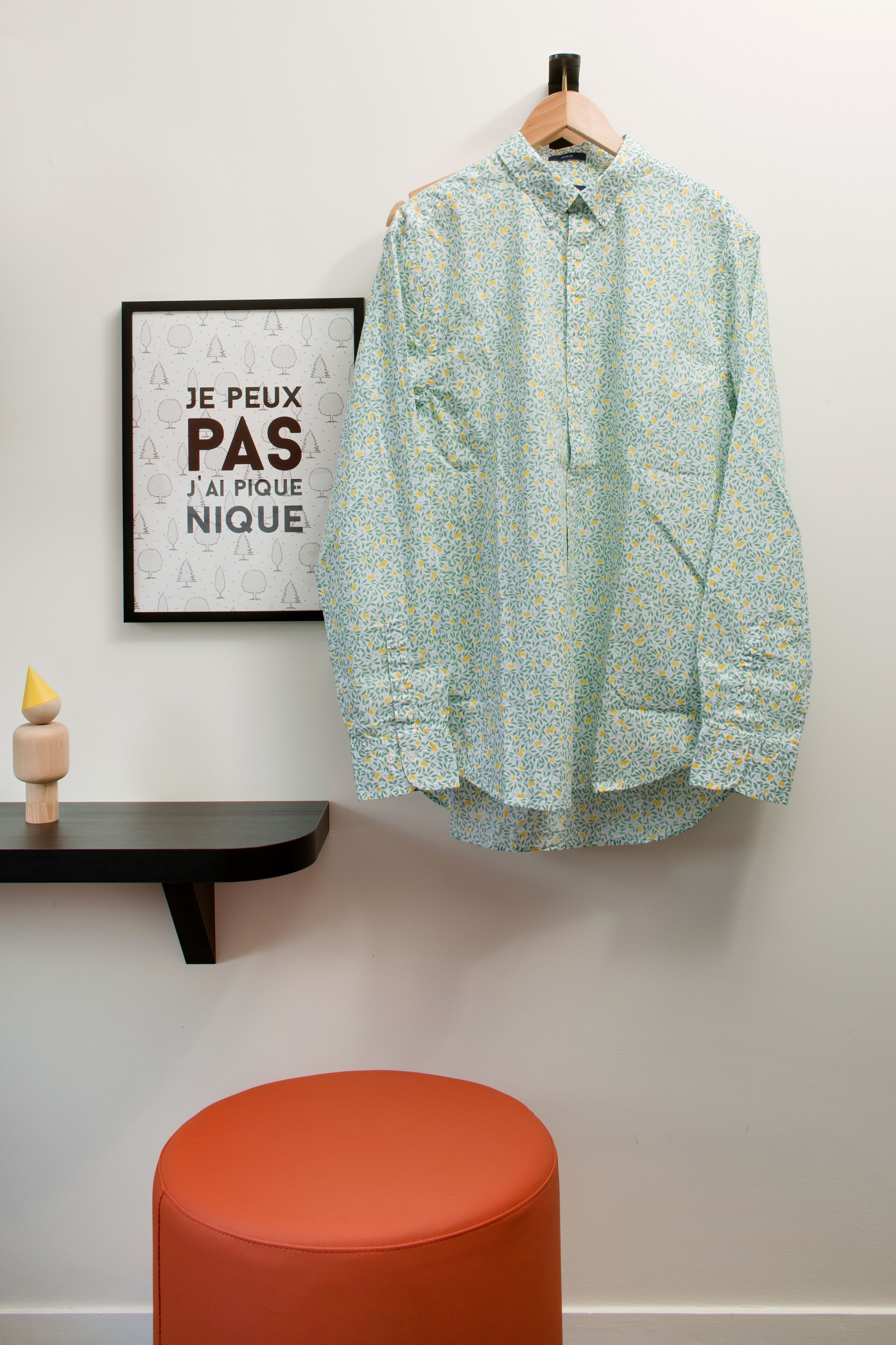 A room with a light blue patterned shirt hanging on a wooden hanger, a framed quote on the wall, a small wooden stool, and an orange ottoman.