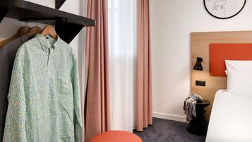 Una habitación de hotel con una cama, una mesita de noche, un reloj de pared, un perchero y una ventana con cortinas.