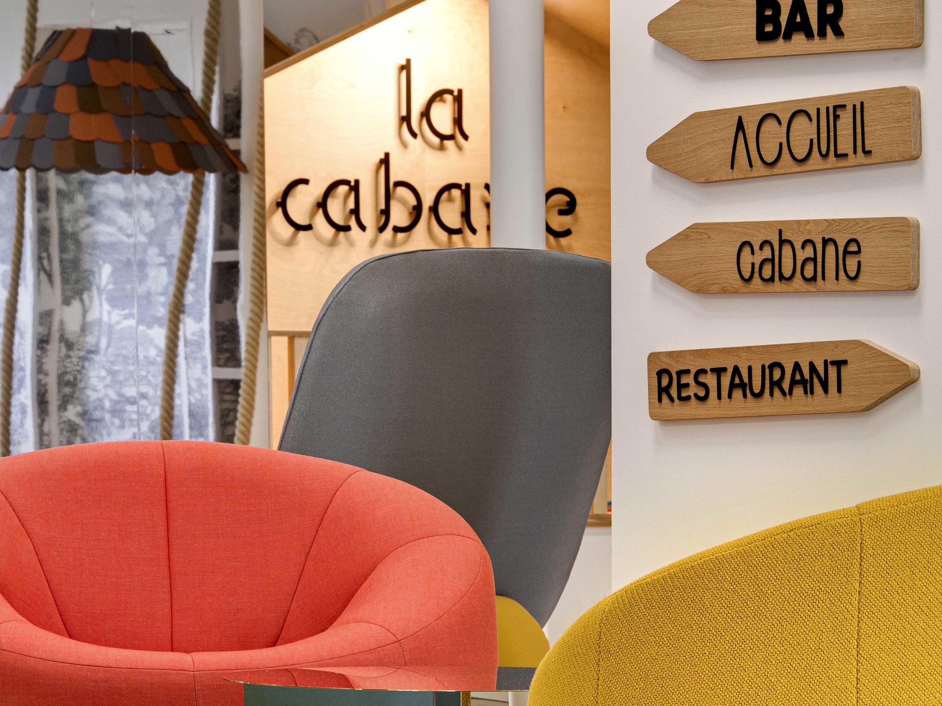 Un interior moderno con una silla roja, una silla gris y una silla amarilla. En la pared, hay letreros de madera que indican un bar, un restaurante y una cabaña.