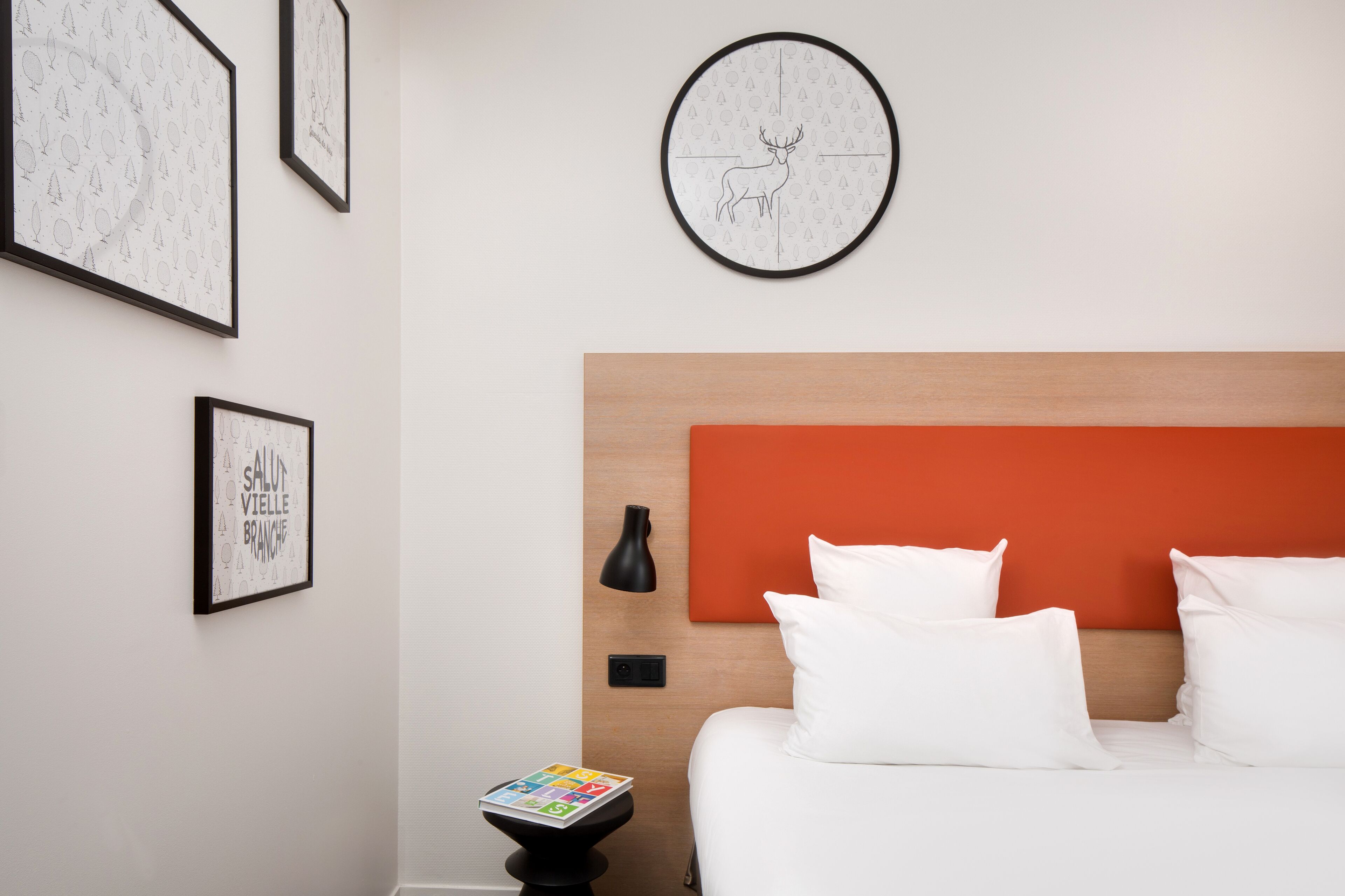Een hotelkamer met een bed, witte kussens, een nachtkastje met een lamp en een kunstwerk in een lijst aan de muur.