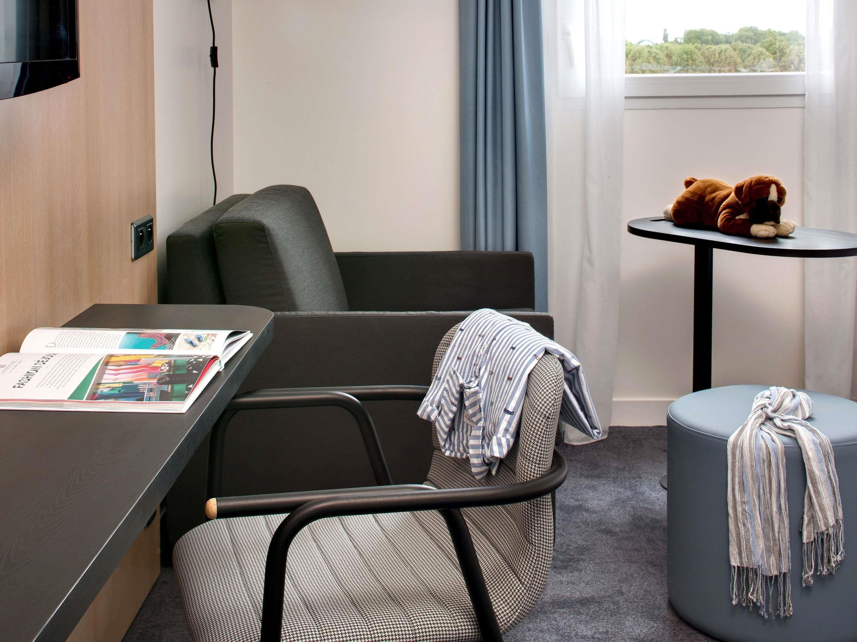 Una habitación de hotel con una cama, un escritorio con una revista, una silla, una mesita redonda con un osito de peluche y una ventana con cortinas.