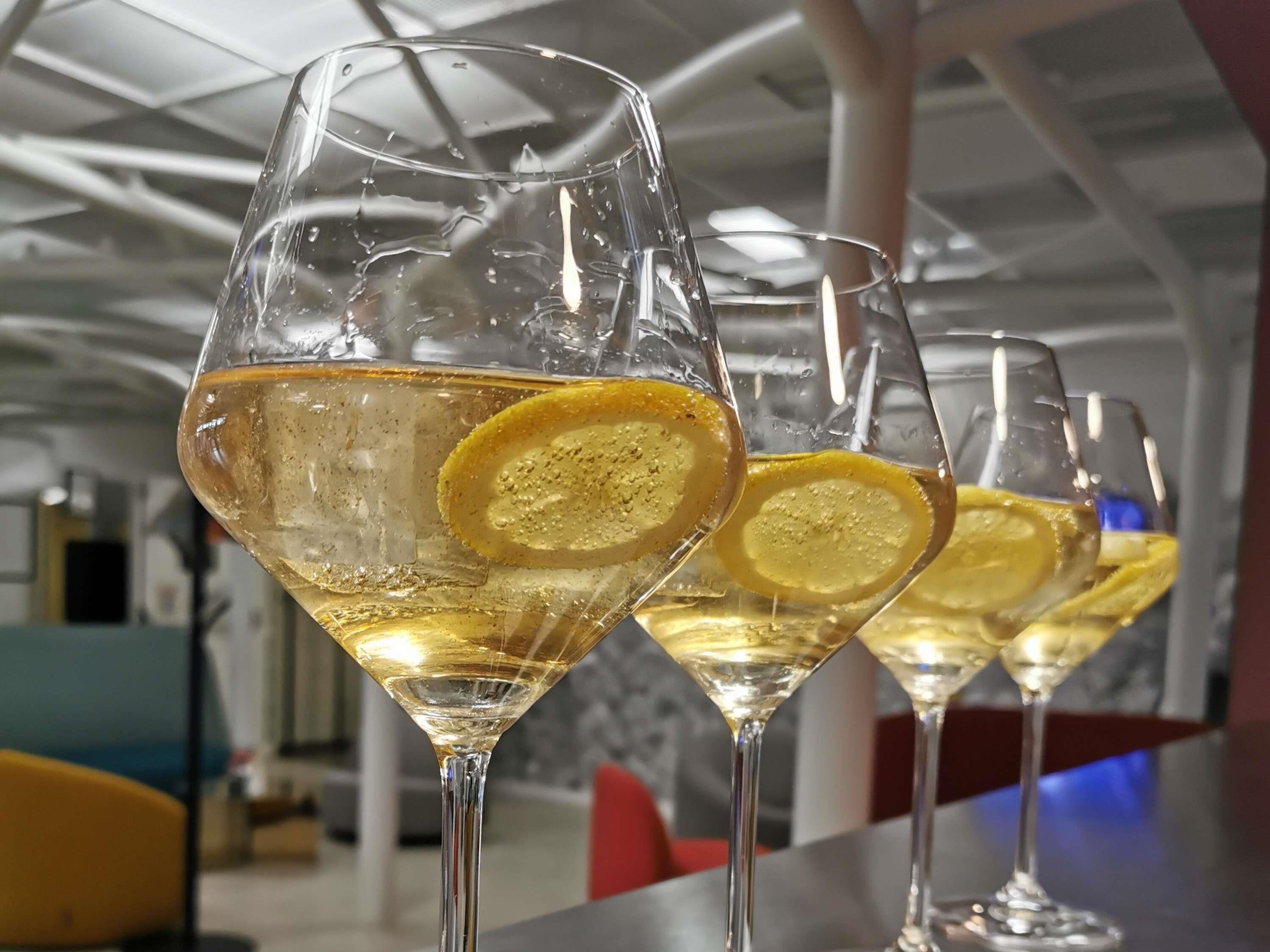 Three glasses of white wine with lemon slices, placed on a reflective surface in a modern interior with a high ceiling and abstract art.