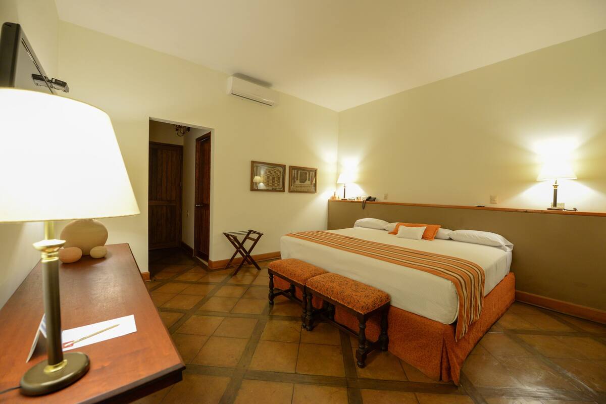 Een hotelkamer met een bed, een bureau met lamp, een bankje en twee wandlampen.