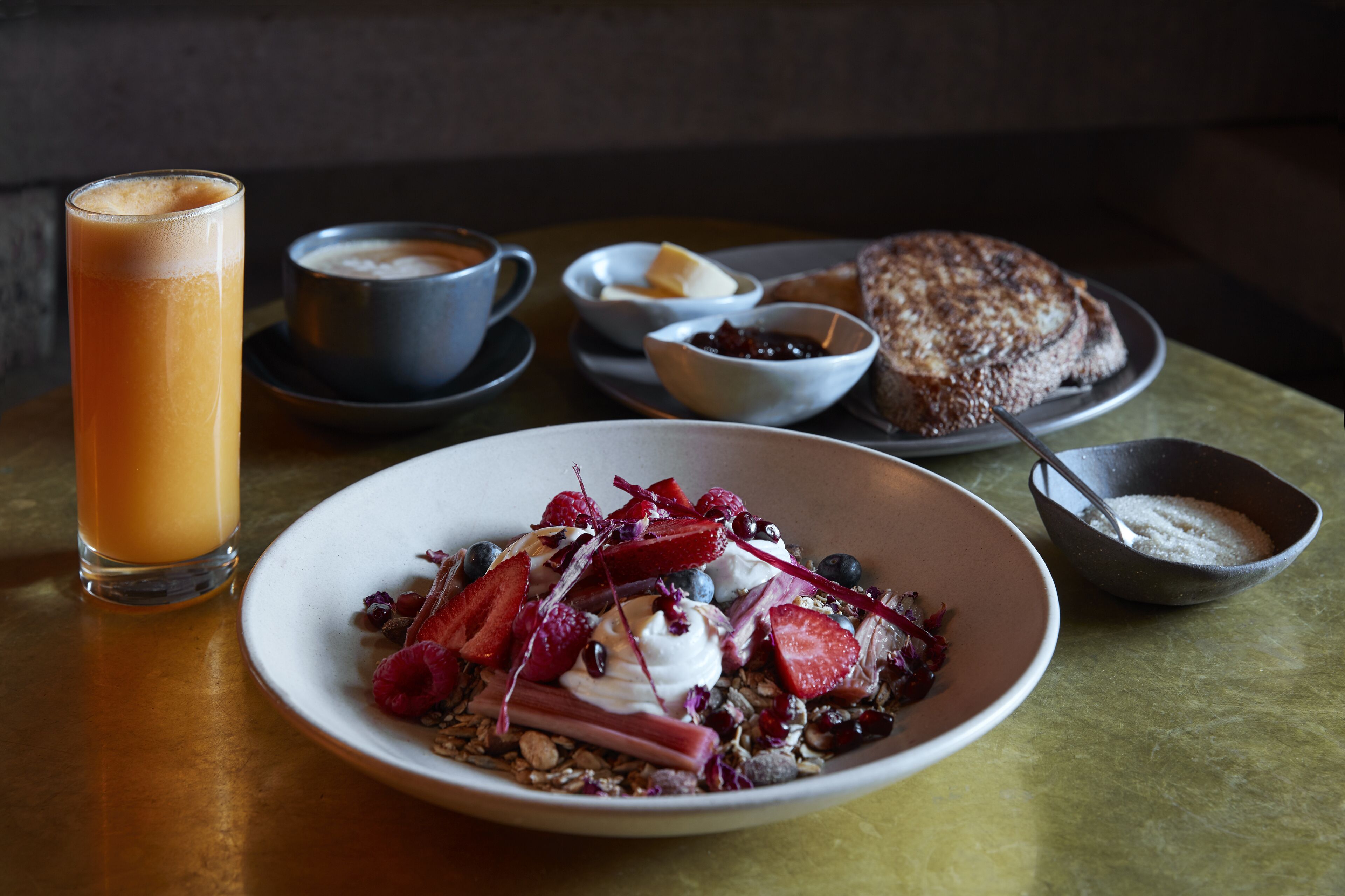 A breakfast setting with a plate of cereal topped with fruits and yogurt, a glass of orange juice, and a slice of bread on a table.