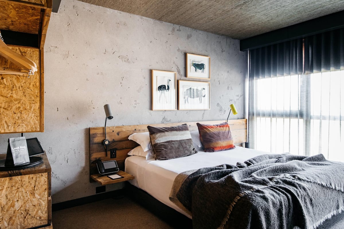 Un dormitorio con cabecera de madera, cama con manta gris, escritorio con ordenador portátil y ventana con cortinas.