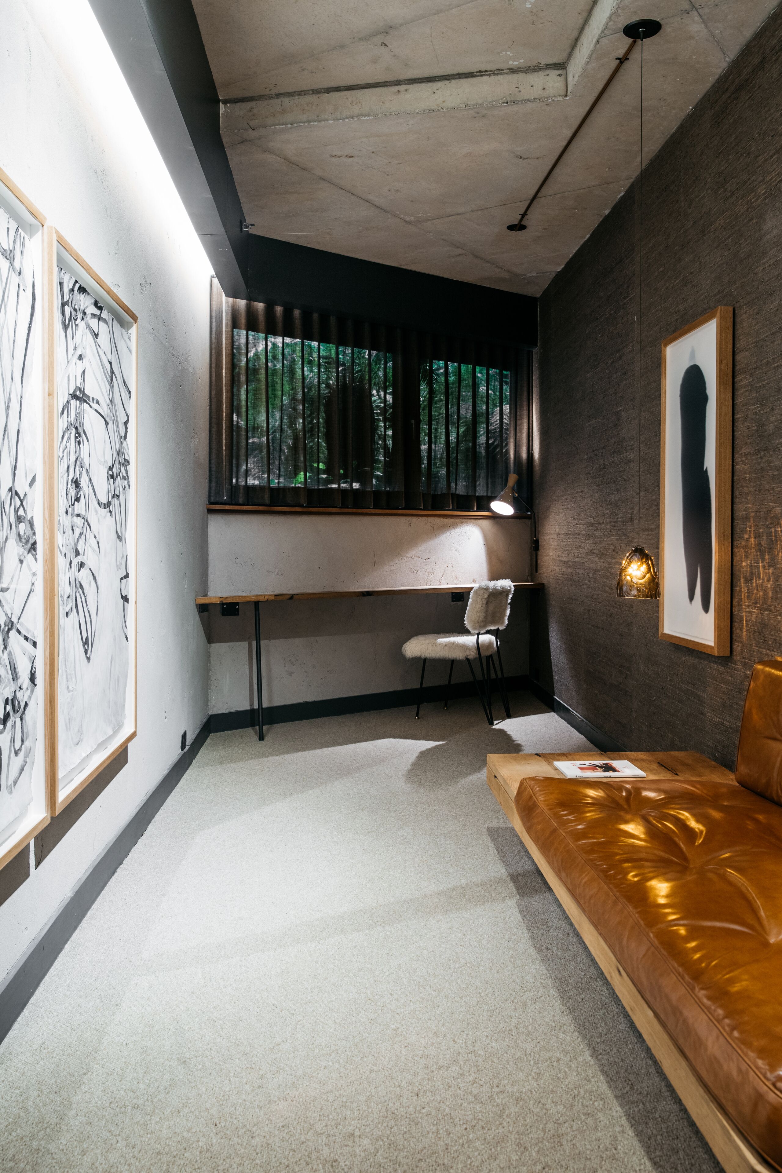 A modern room with a desk, chair, and sofa. There is a large window with blinds, a ceiling light, and artwork on the walls.