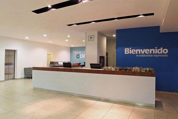A reception area with a blue wall, a wooden reception desk, and a sign that reads 'Bienvenido te estamos esperando'. - One Querétaro Aeropuerto Hotel (El Marques)