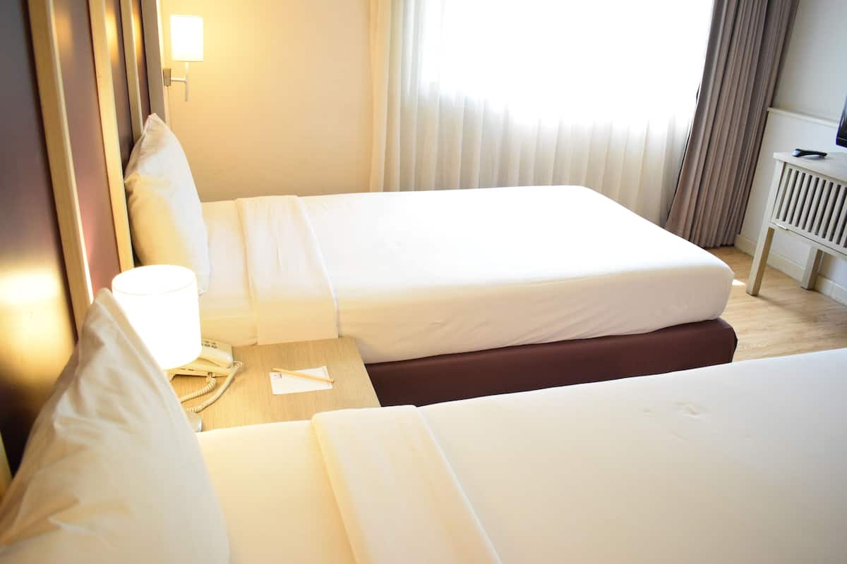 Une chambre d’hôtel avec deux lits, une table de chevet avec une lampe, un téléphone et une fenêtre avec des rideaux.