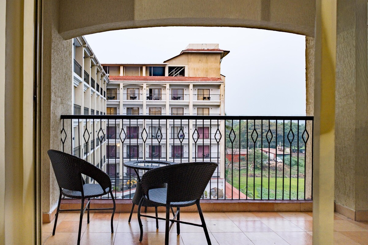 Ein Balkon mit zwei Stühlen und einem Tisch, der auf ein Gebäude mit mehreren Balkonen und eine grüne Landschaft blickt.
