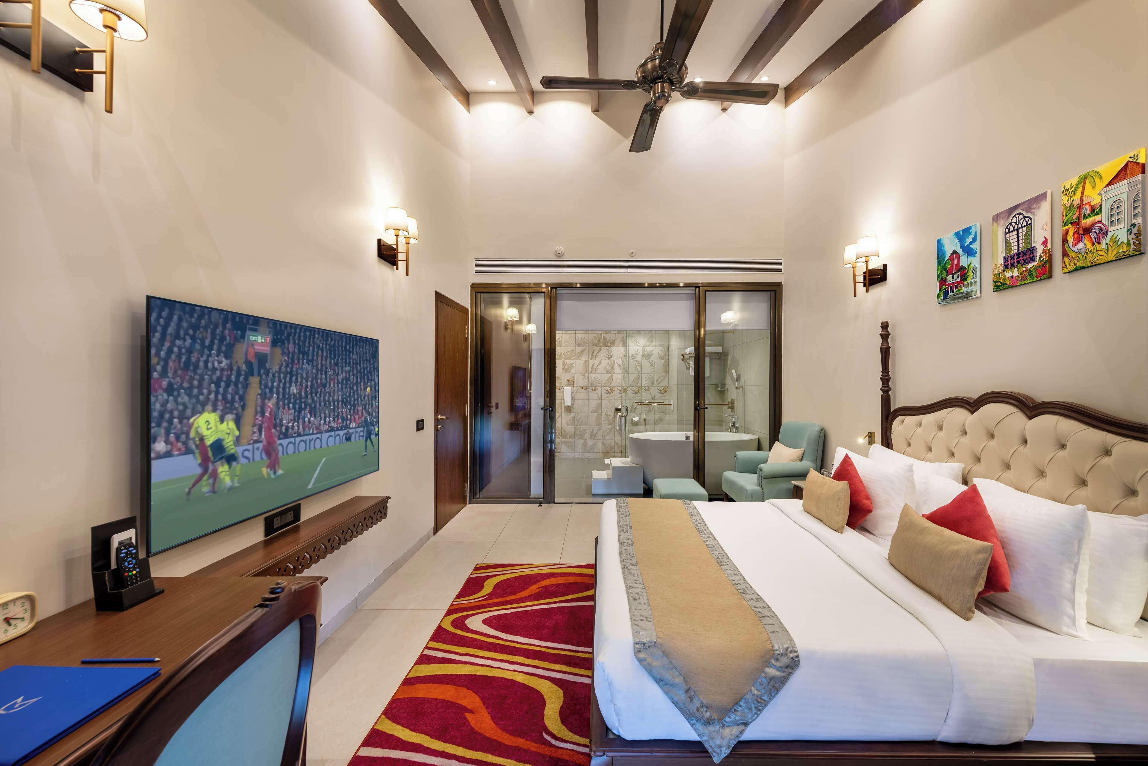 A hotel room with a large bed, a television, a desk with a computer, and a bathroom visible through a glass door.