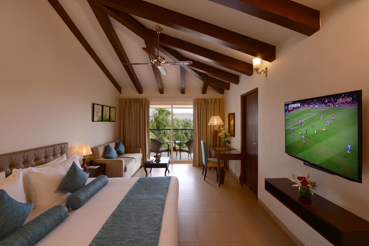 Ein Hotelzimmer mit einem großen Bett, einem Fernseher, der ein Fußballspiel zeigt, einem Balkon mit Blick und einem Deckenventilator.
