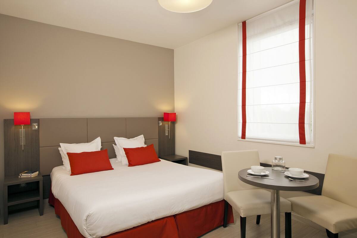 Une chambre d’hôtel comprenant un lit, deux tables de chevet, une table à manger ronde avec des chaises et une fenêtre avec des rideaux.