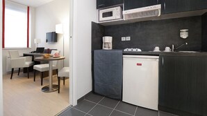 Cocina compacta equipada con microondas, cafetera y un refrigerador pequeño. Cuenta con un comedor con mesa y sillas, y un televisor montado en la pared.