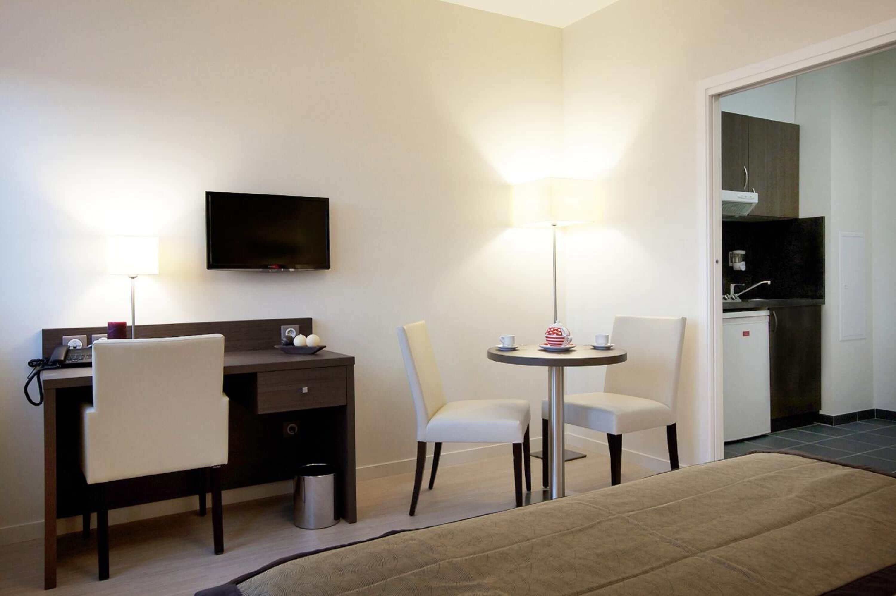 Habitación de hotel con escritorio, dos sillas, televisor, lámpara, una mesita con un pastel y una kitchenette al fondo.
