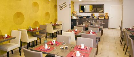 A dining area with tables set for breakfast, a kitchen in the background, and a clock on the wall.