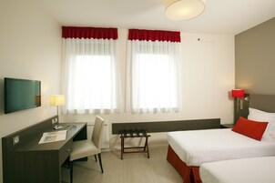Ett hotellrum med två sängar, ett skrivbord, en stol, en tv och ett fönster med gardiner.
