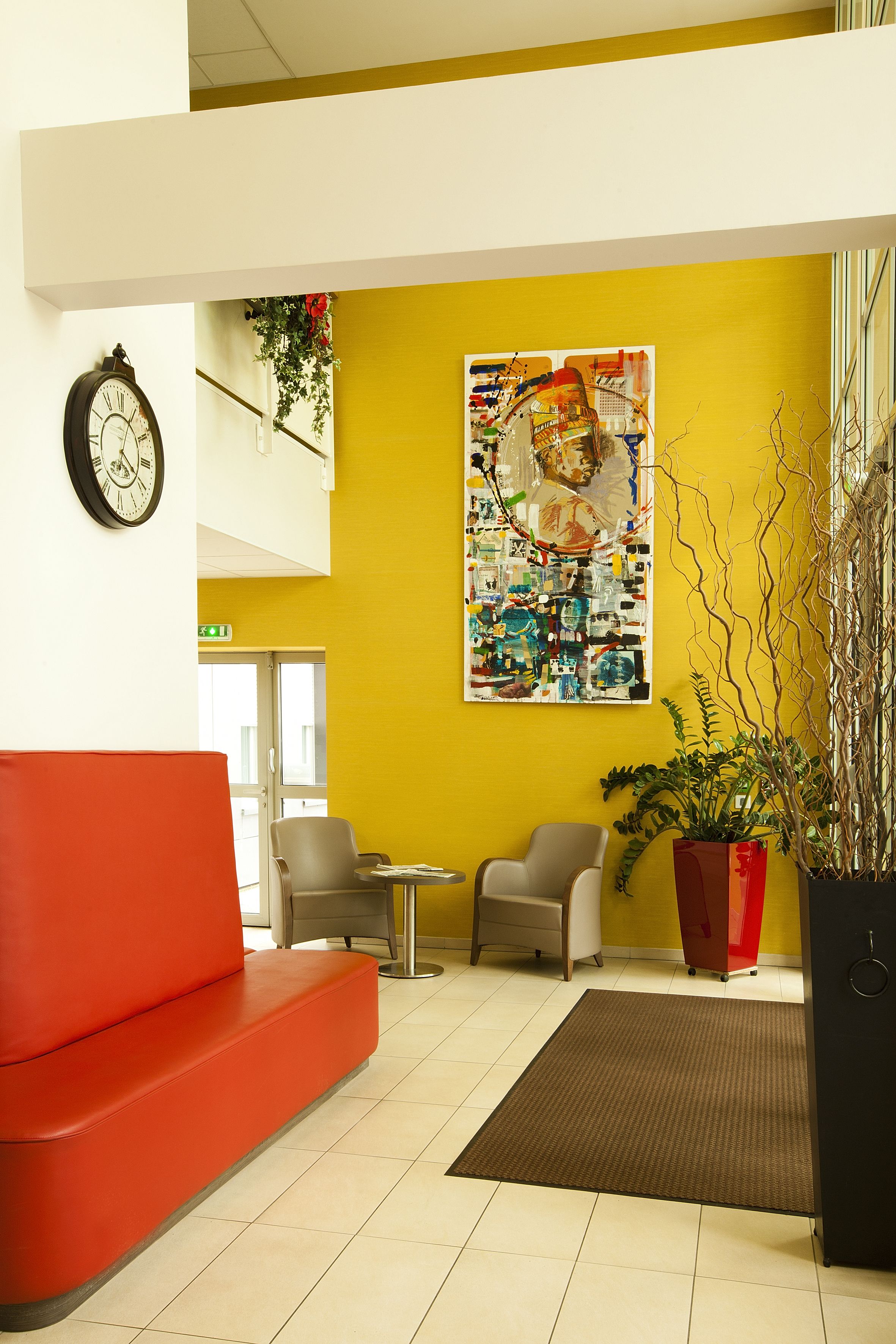 Un hall d’entrée avec un canapé rouge, deux fauteuils, une grande peinture abstraite, une horloge et une plante en pot.