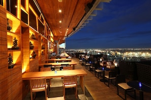 Un restaurante en la azotea con mesas y sillas de madera, vistas a la ciudad y estantes con adornos.