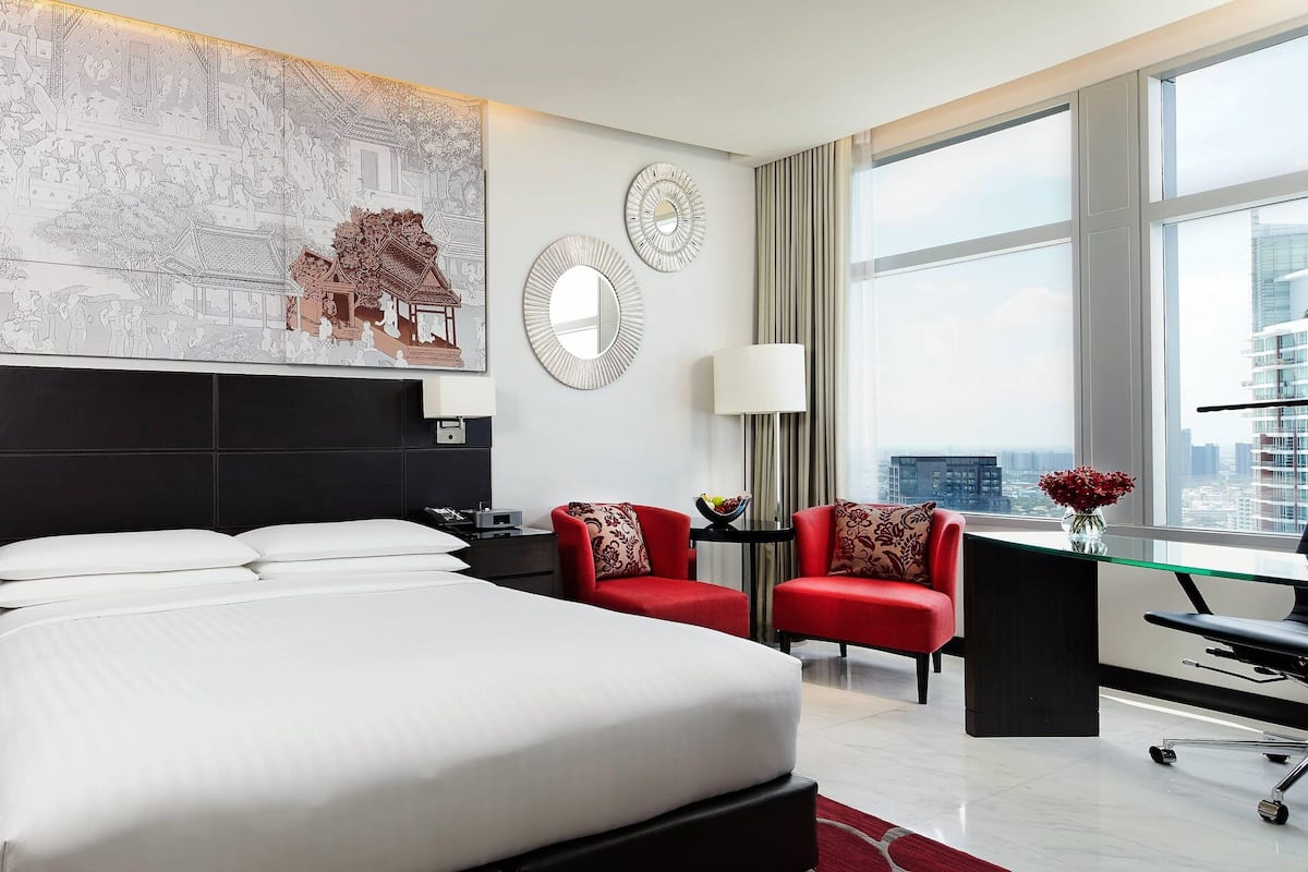 Una habitación de hotel con una cama grande, dos sillones rojos, un escritorio con una silla, un ventanal con vistas a edificios y una obra de arte en la pared.