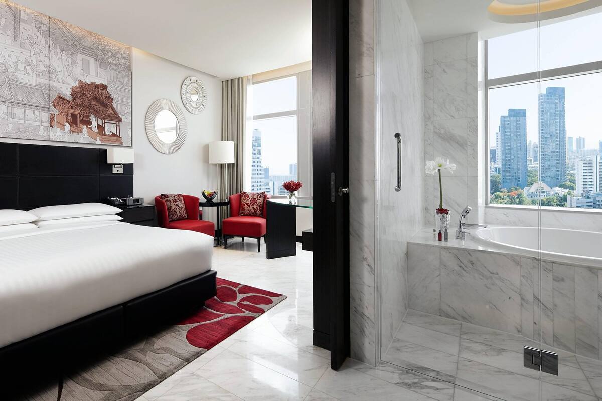Una habitación de hotel moderna con una cama grande, un sofá rojo, un escritorio y un baño con ventana amplia y vista a la ciudad.