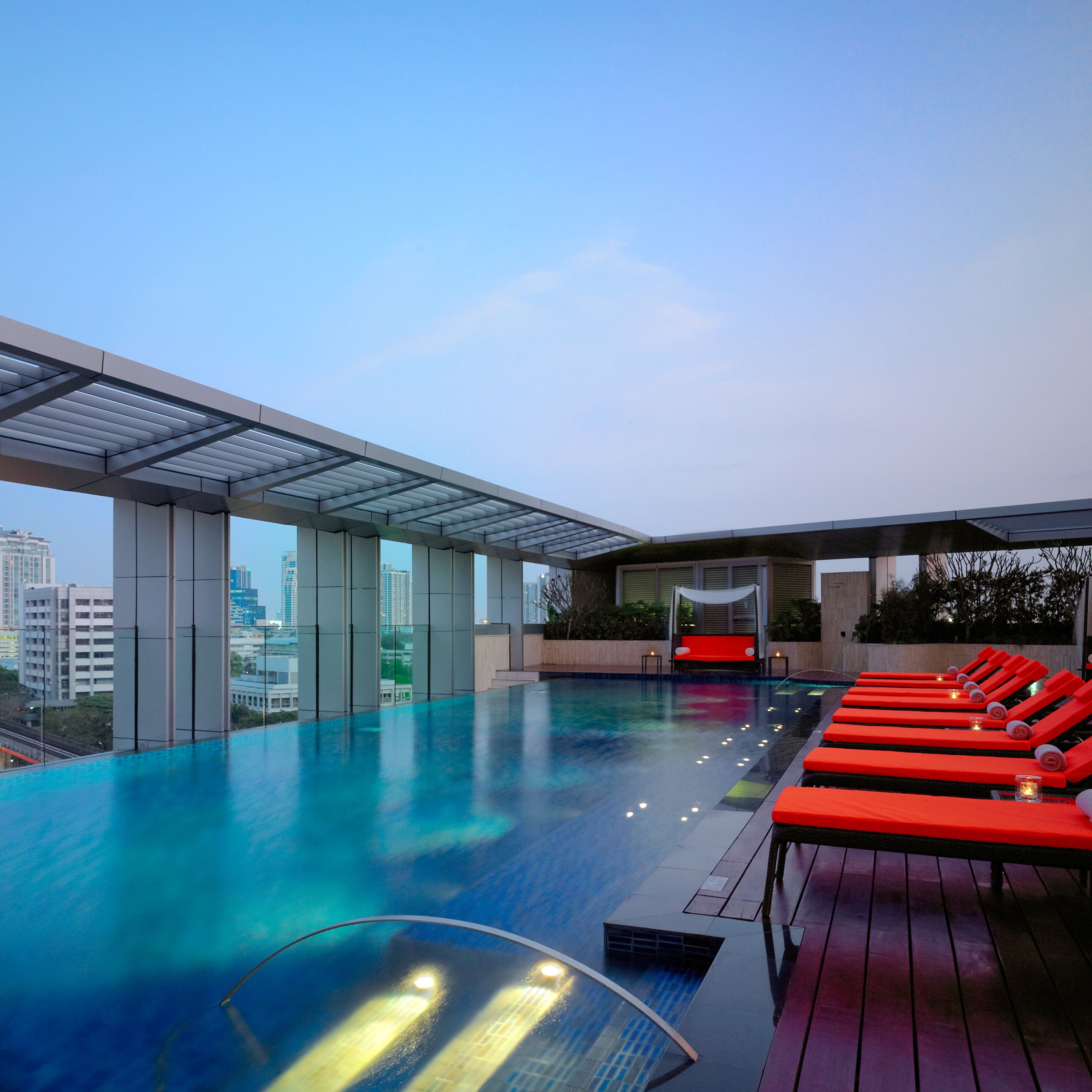A rooftop infinity pool with red lounge chairs and a glass balustrade.