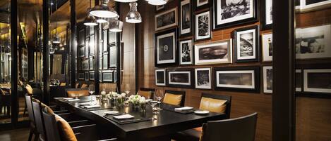 A dining area with a long table set for six, surrounded by chairs. The walls are adorned with framed photographs.