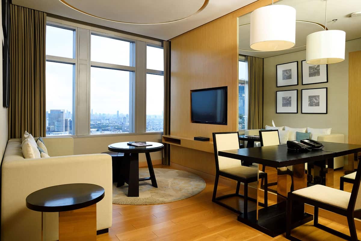 Una habitación de hotel moderna con un gran ventanal que ofrece vistas a la ciudad, un televisor de pantalla plana montado en la pared, una zona de comedor con mesa y sillas, y un sofá.