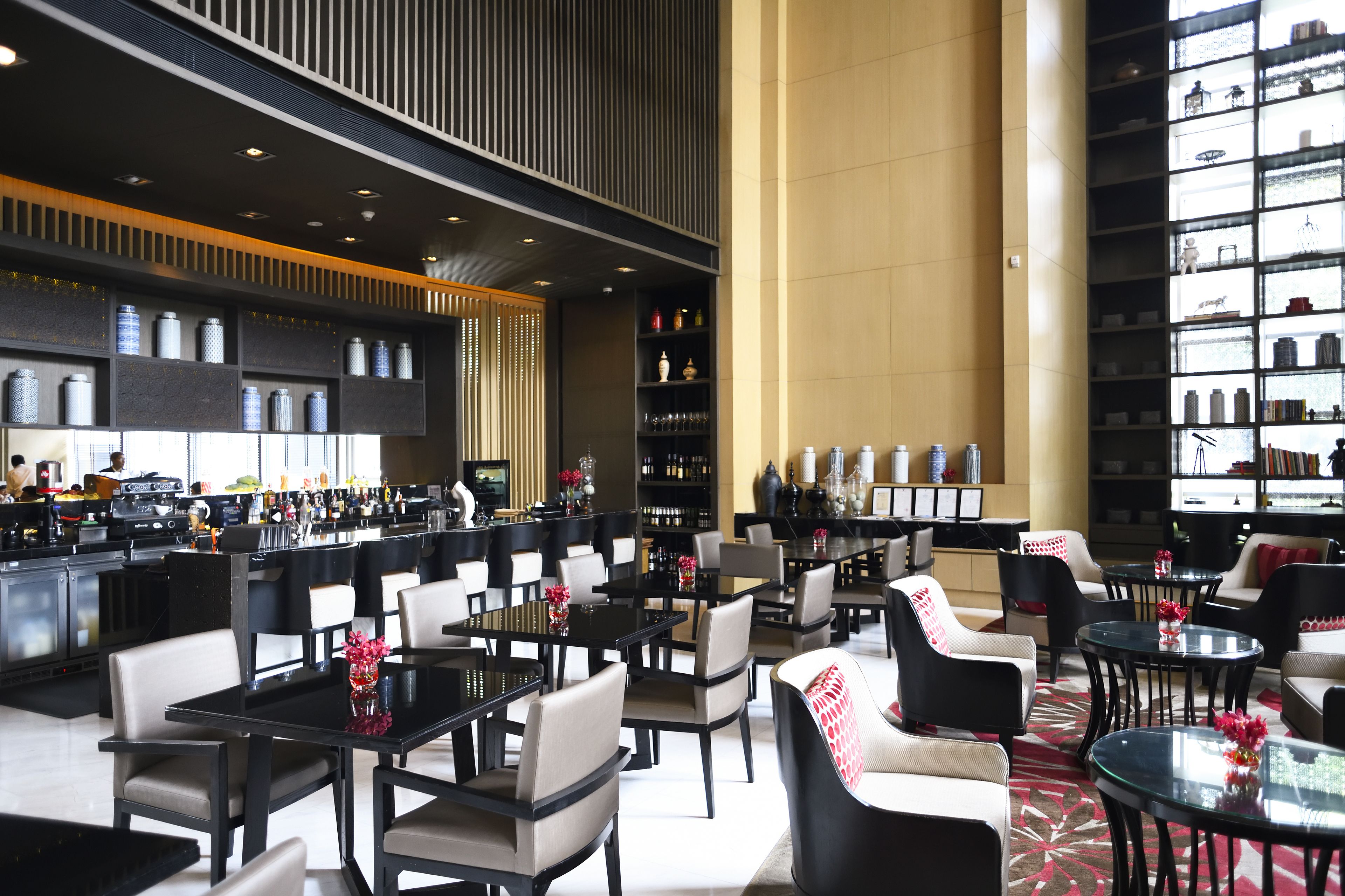 A modern hotel lobby with multiple dining tables and chairs, a bar area, and a large bookshelf filled with books and decorative items.