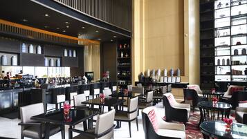 A modern hotel lobby with multiple dining tables and chairs, a bar area, and a large bookshelf filled with books and decorative items.