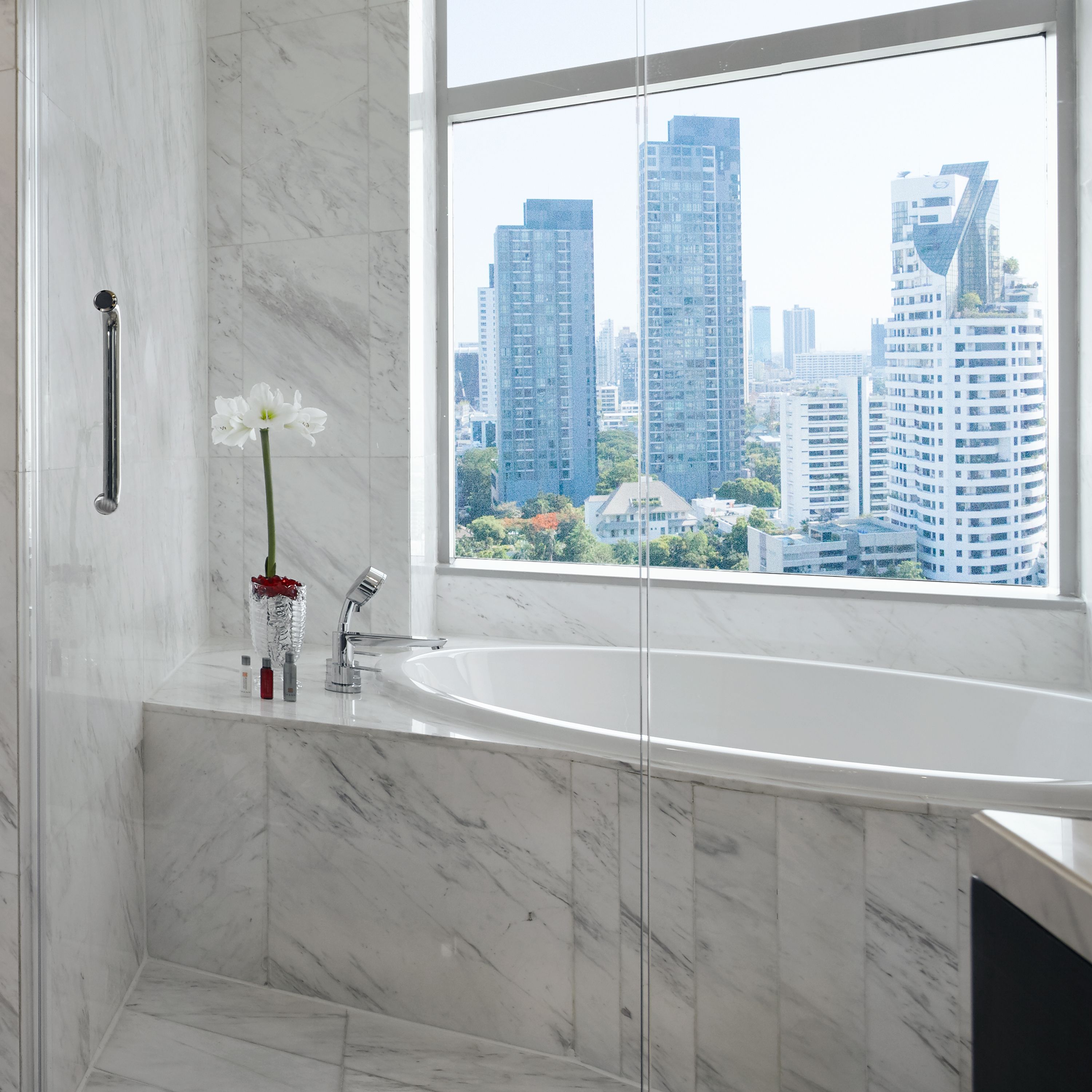 A modern bathroom with a marble sink, a glass shower enclosure, and a view of tall buildings through a large window.