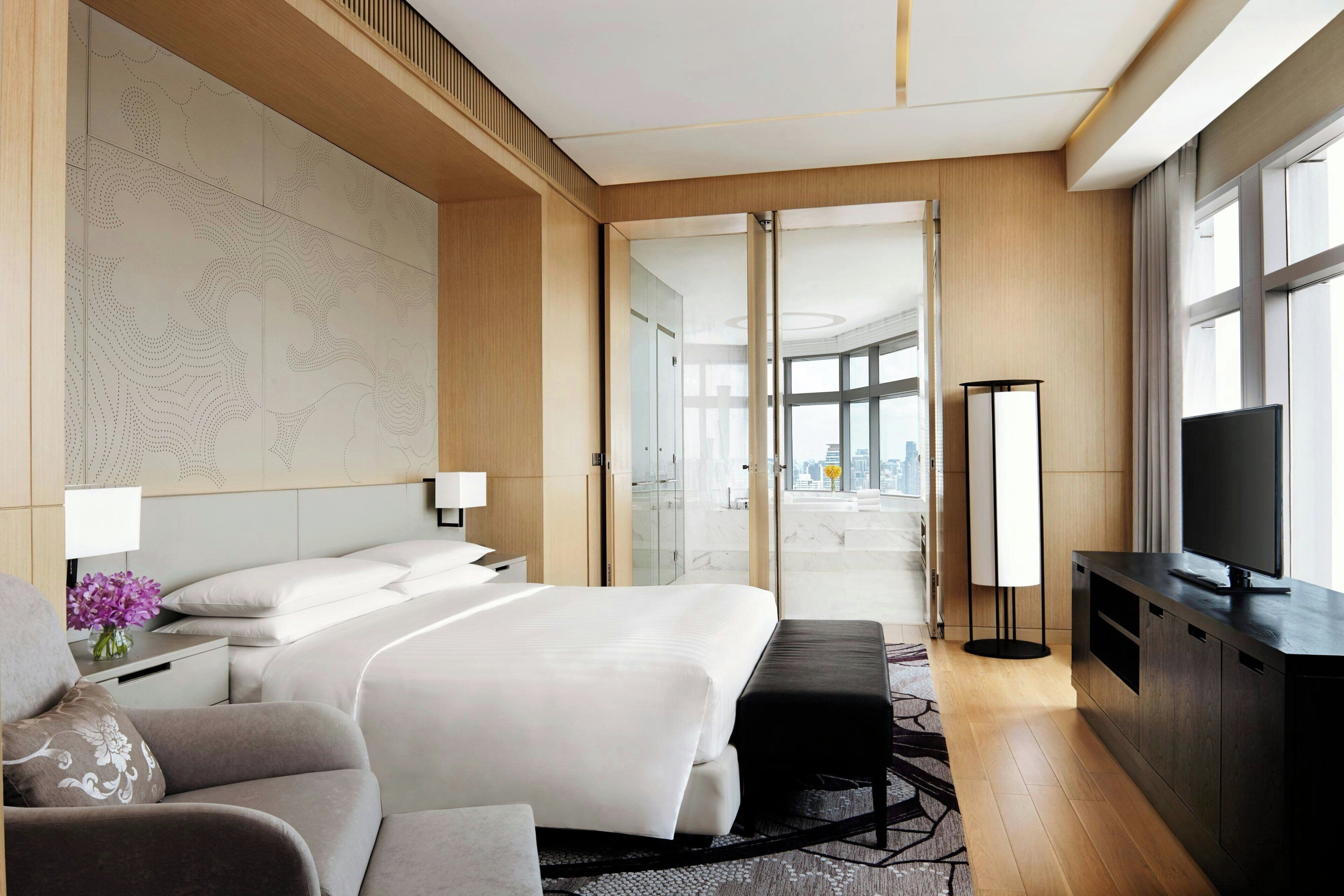 Habitación de hotel moderna con una cama grande, un sofá, televisión de pantalla plana y vistas a la ciudad a través de amplios ventanales.