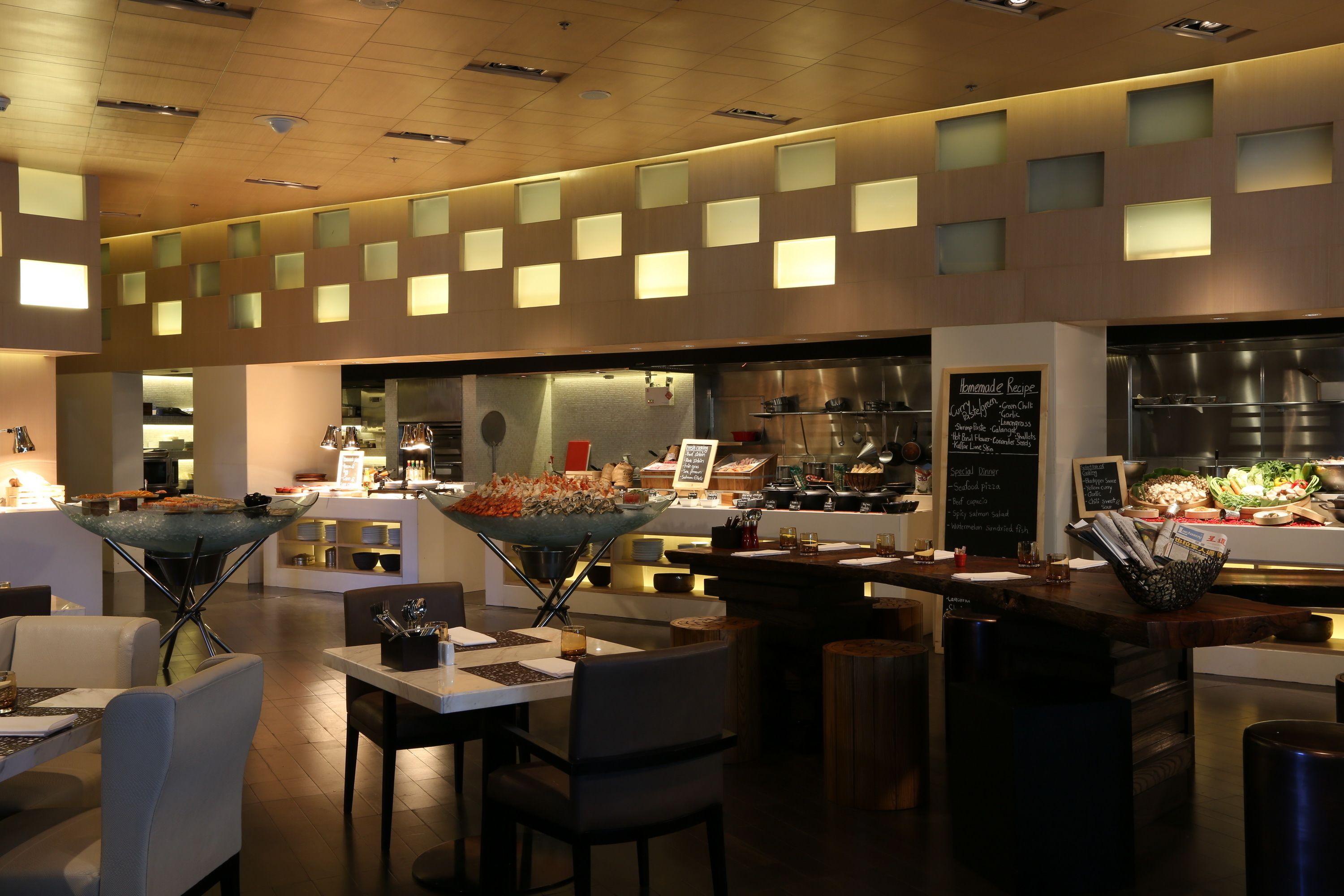 A modern restaurant interior with a central serving area, multiple tables, and a visible kitchen in the background.