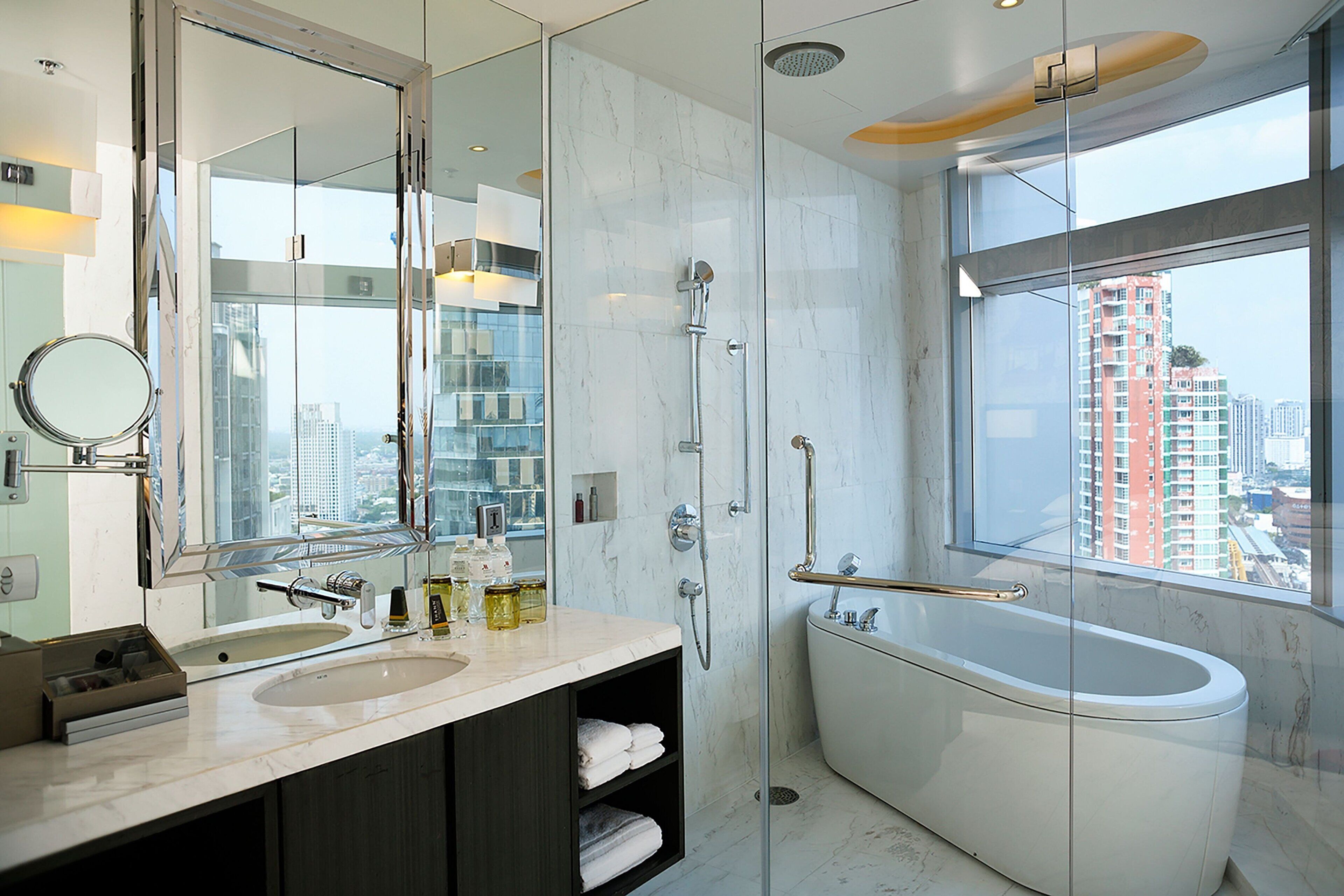A modern bathroom with a large mirror, two sinks, a bathtub, and a window offering a city view.