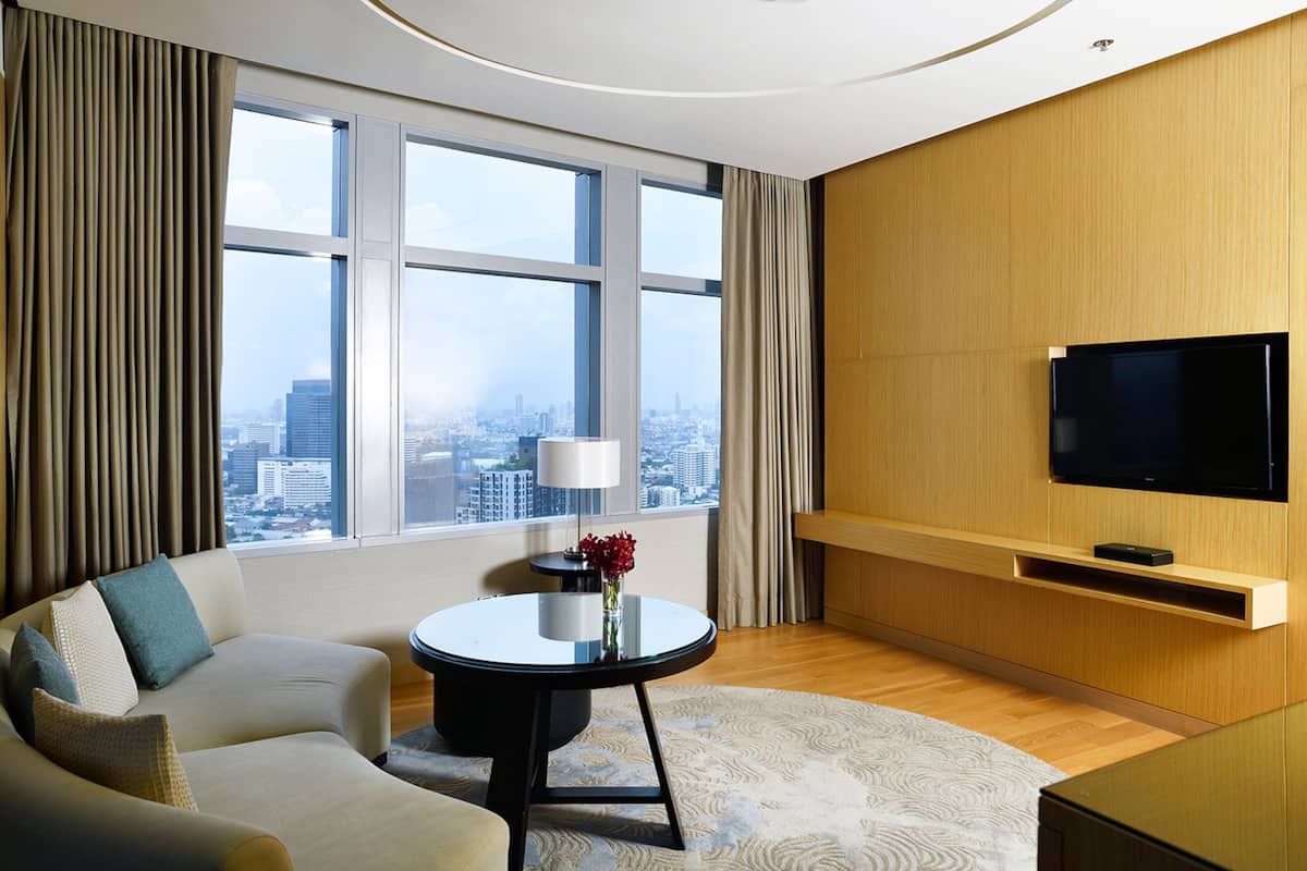 Habitación de hotel moderna con un ventanal grande que ofrece vistas a la ciudad, un sofá, una mesita redonda con una lámpara y un televisor montado.