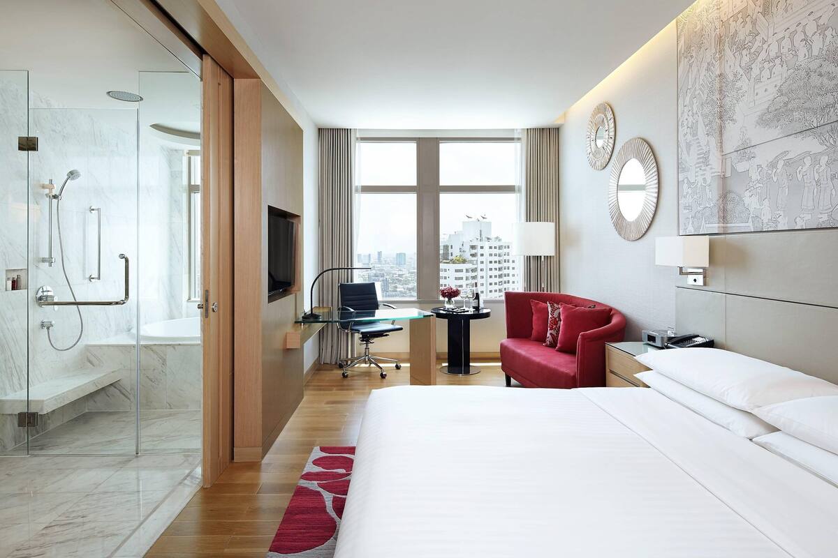 Habitación de hotel con cama, escritorio, una silla roja, televisor, ducha y una ventana grande con vistas a la ciudad.