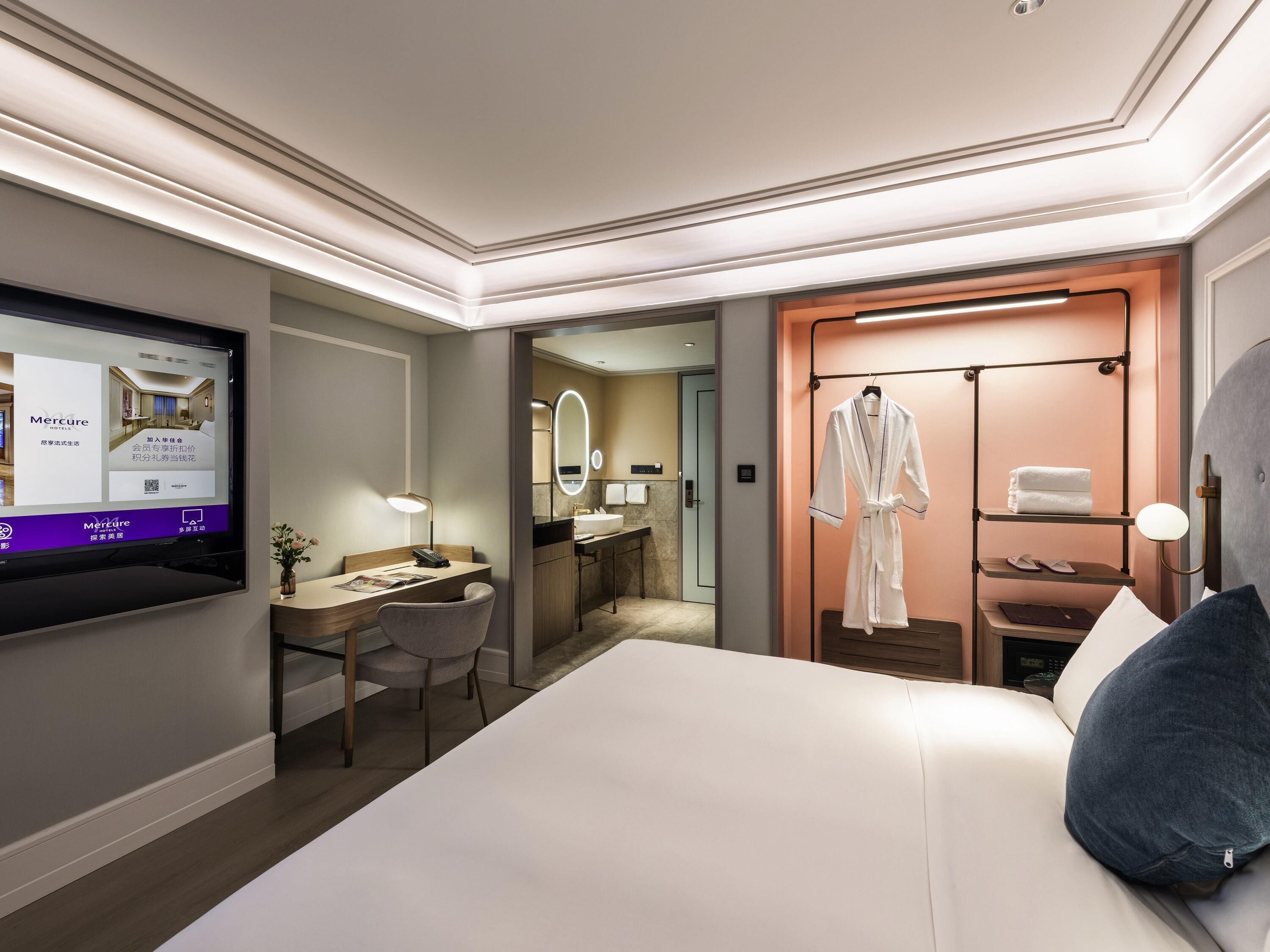 A hotel room with a bed, desk, chair, robe rack, and a TV displaying hotel information.
