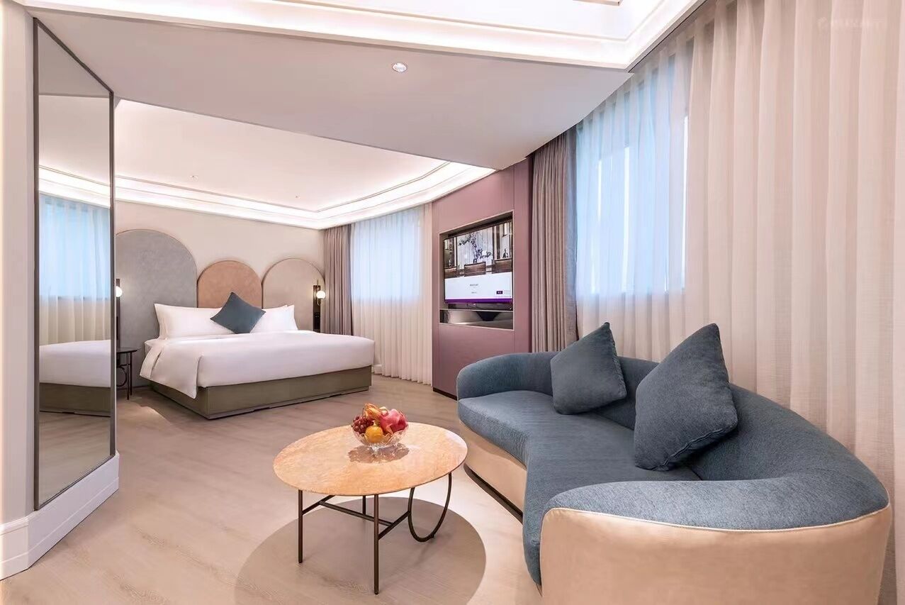 A modern hotel room with a bed, a sofa, a TV, and a small table with fruit.