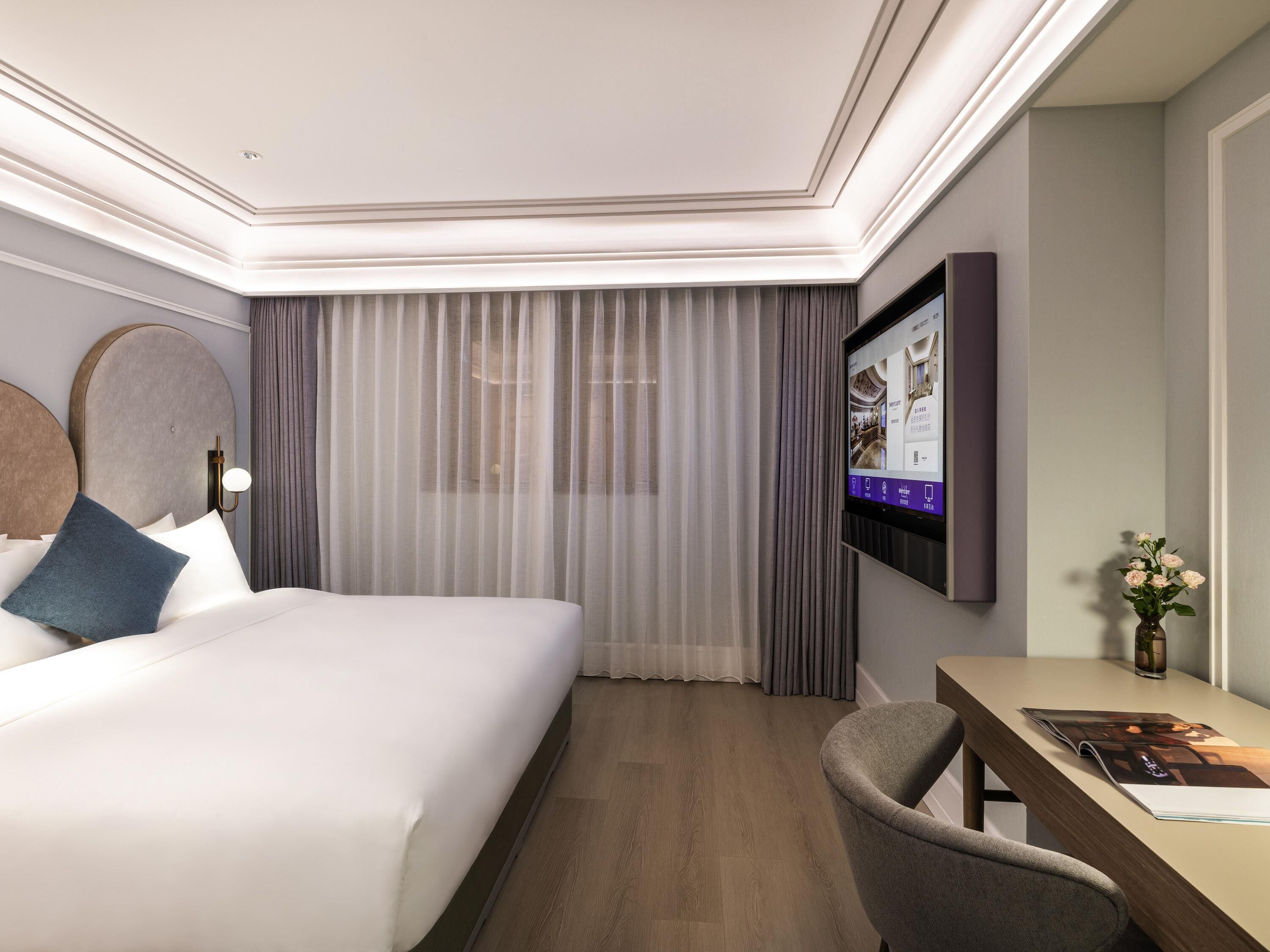 Quarto de hotel com uma cama grande, uma escrivaninha, uma cadeira, uma televisão e cortinas.