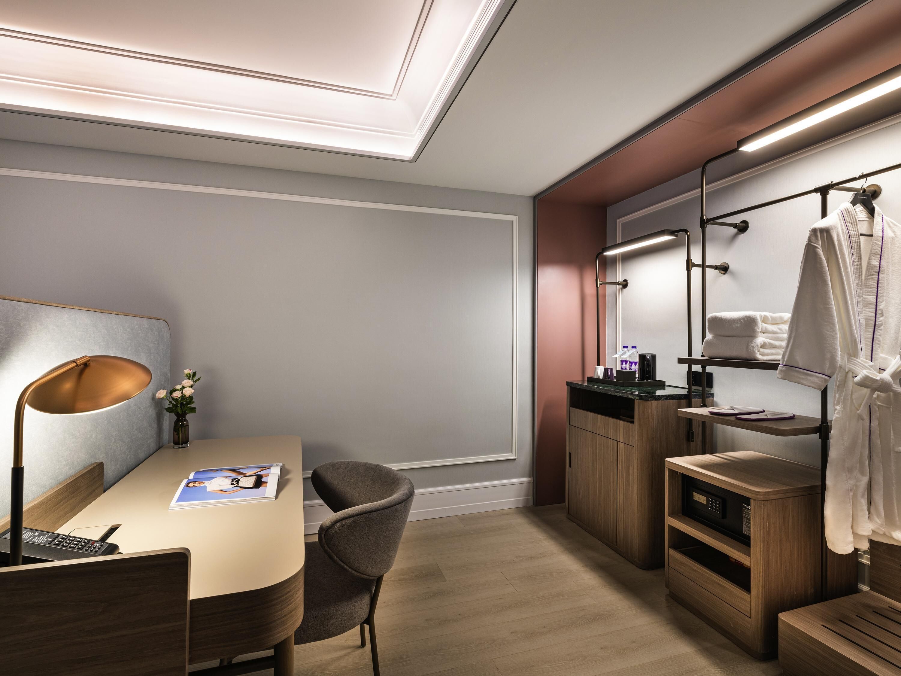 A modern hotel room with a desk, chair, bedside table, and a mirror with a robe hanging.