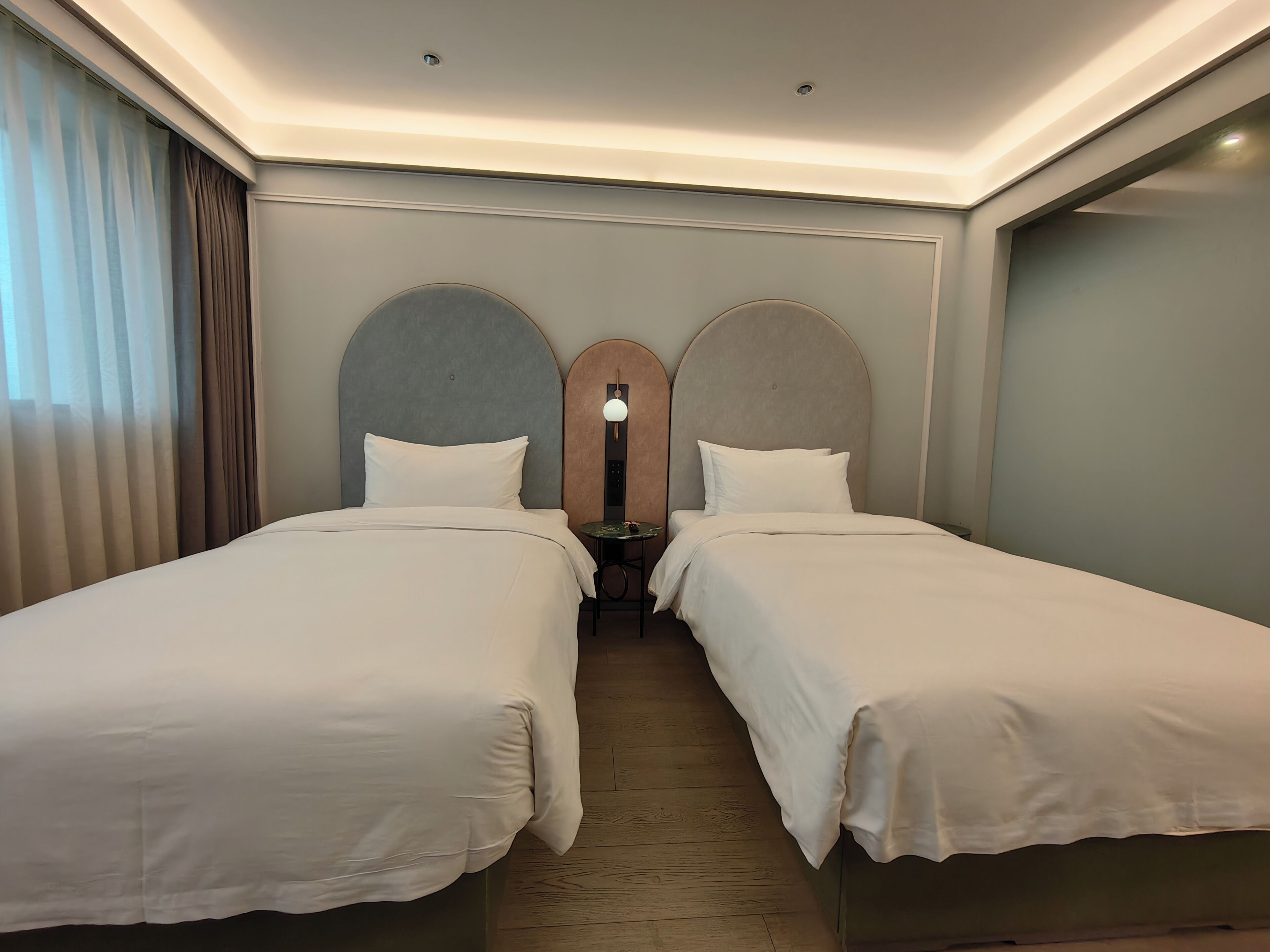 A hotel room with two beds, a wooden floor, a nightstand, and a wall-mounted light fixture.