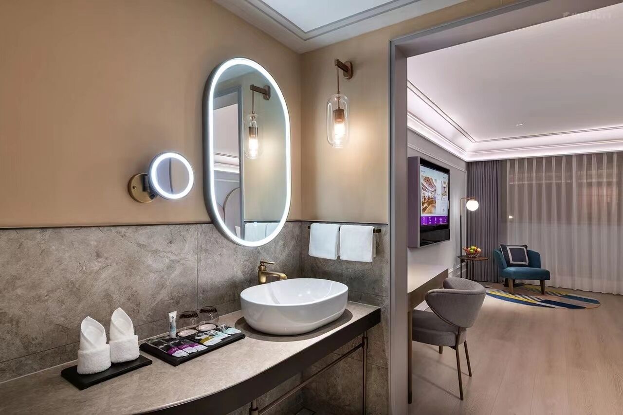 Un baño moderno con un lavamanos ovalado grande, un espejo redondo con luz y un televisor de pantalla plana.