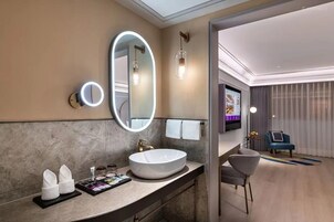 Un baño moderno con un lavamanos ovalado grande, un espejo redondo con luz y un televisor de pantalla plana.
