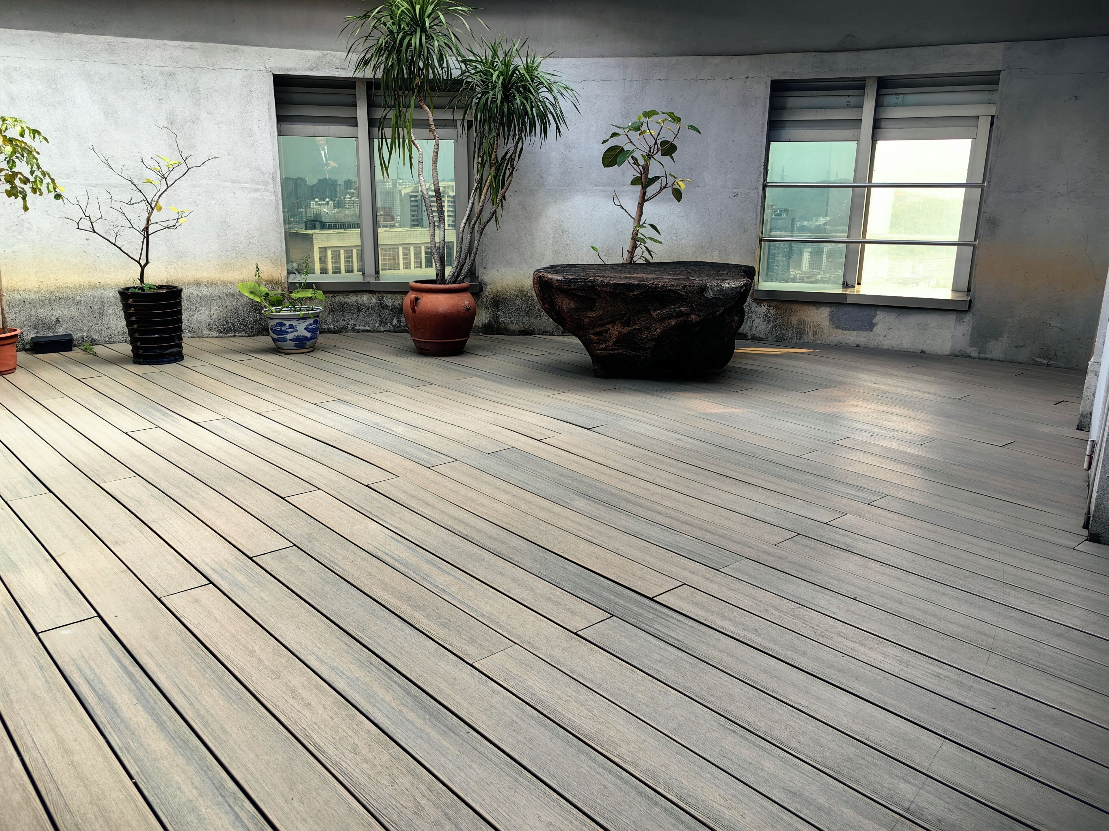 Une terrasse sur le toit, dotée d’un plancher en bois, de plantes en pot et de grandes fenêtres offrant une vue sur le paysage urbain.
