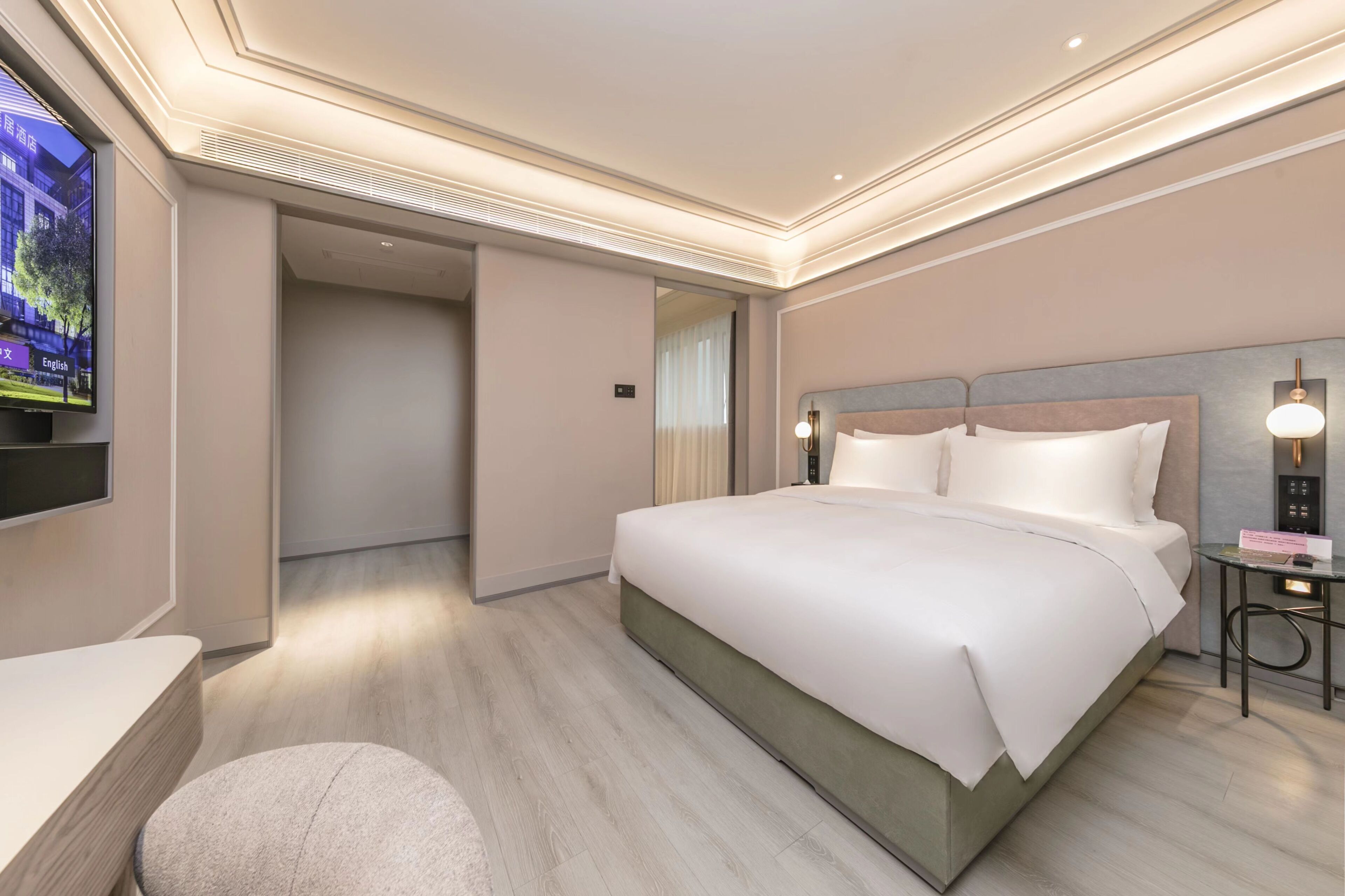 A modern hotel room with a large bed, a TV mounted on the wall, a bedside table, and a small desk with a lamp.