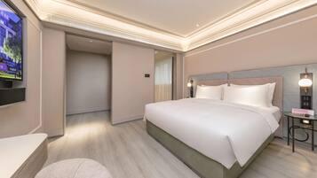A modern hotel room with a large bed, a TV mounted on the wall, a bedside table, and a small desk with a lamp.